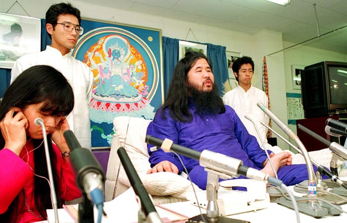 A Japanese man speaks into multiple microphones, flanked by two men and a woman