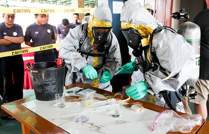 Two people wearing hazmat suits and breathing masks demonstrating handling chemicals