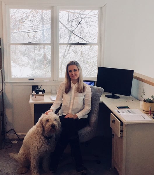A female sitting at a desk in a home office with a dog by her side