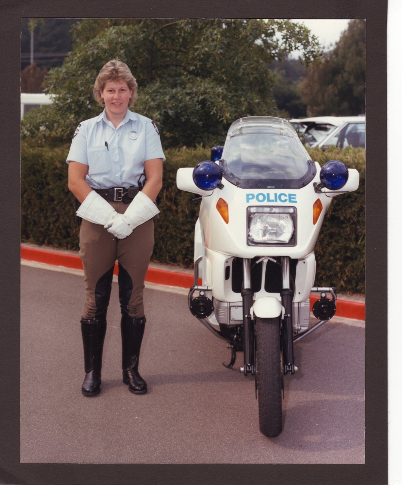 Sergeant  Louise Denley in 1989, standing next to her police motorcycle – black boots, white gloves, riding jodhpurs. 