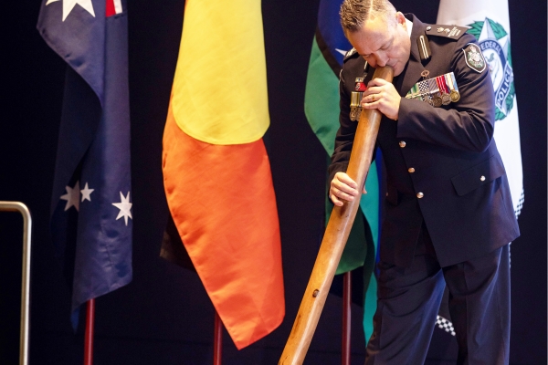 AFP man in ceremonial uniform playing the didgeridoo in front  of an Australian, Aboriginal, Torres Strait Islands, and AFP flag