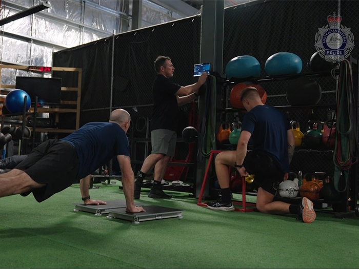 Three people in a gym, one standing, one kneeling, and one in a push up position