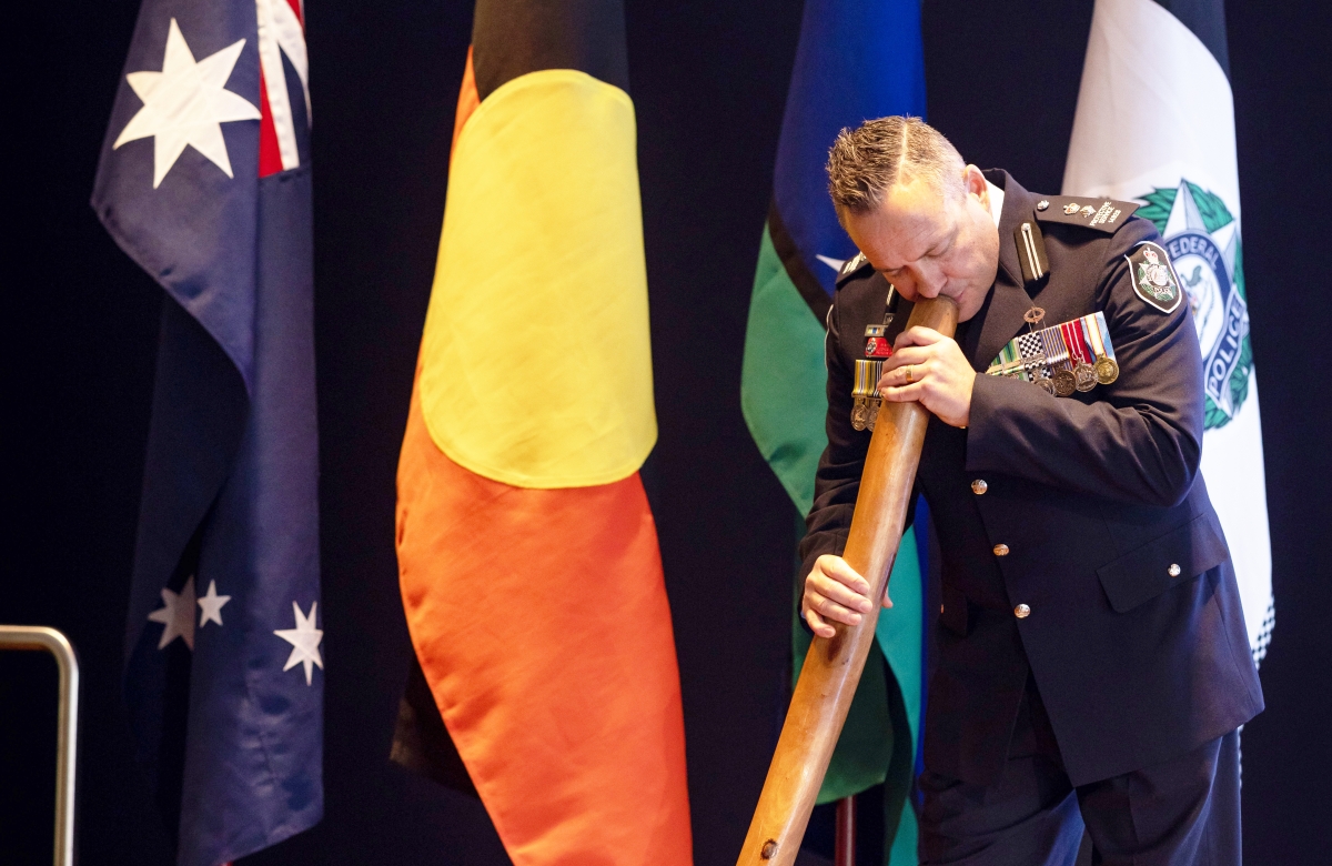 Man in Police ceremonial uniform playing a didgeridoo in front of an Australian, Aboriginal, and AFP flag