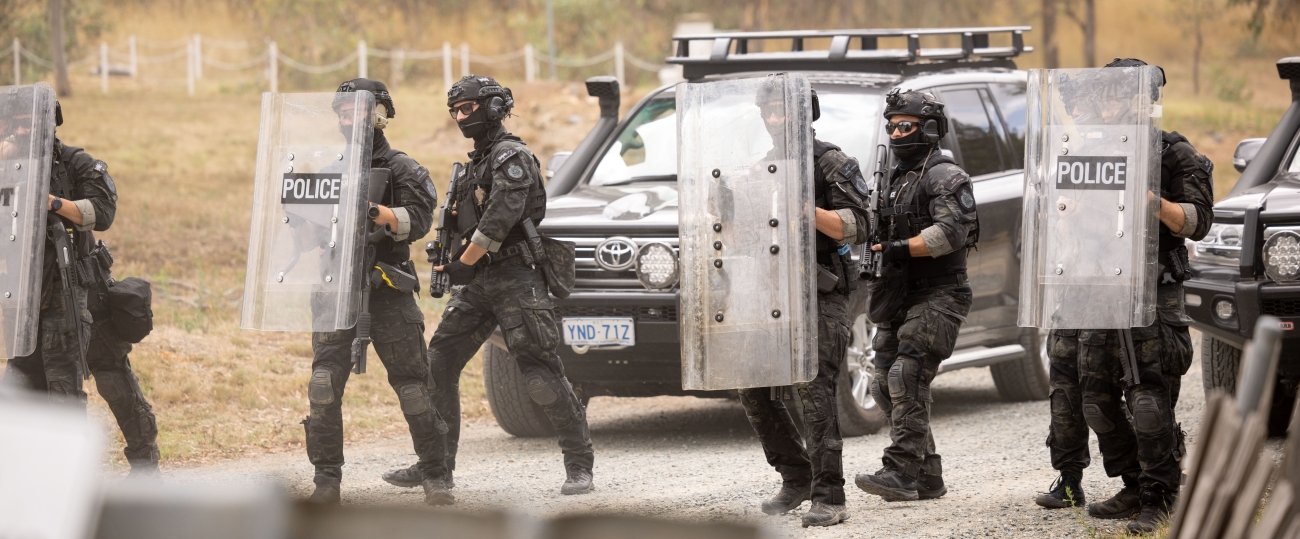 Six armed tactical response police officers advancing in front of a large vehicle